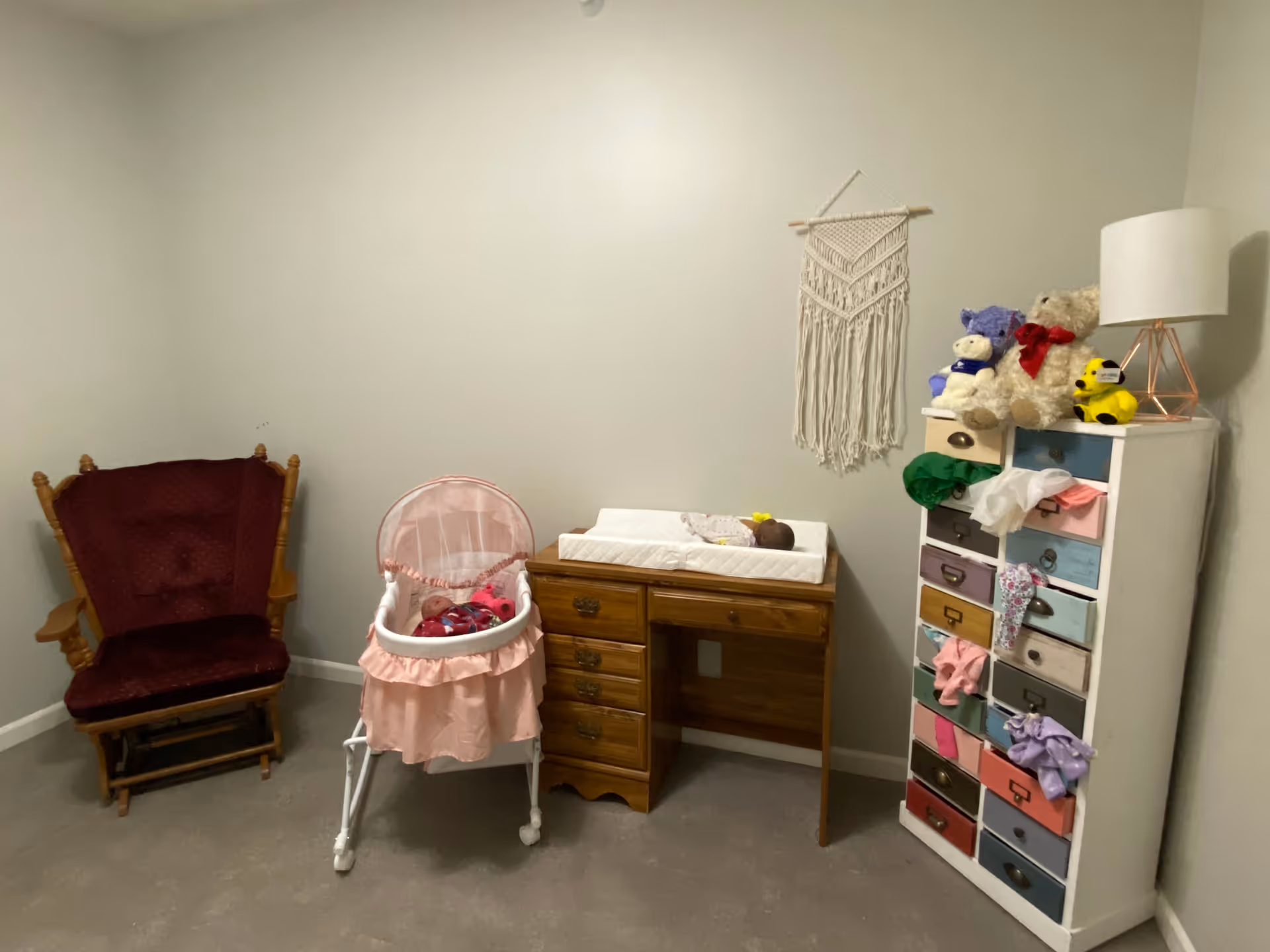 A small room with a wooden rocking chair with maroon cushions on the left, a baby bassinet with a doll inside in the center, a wooden changing table with a doll lying on a changing pad, and a tall white dresser with colorful drawers and stuffed animals on top. A white lamp with a geometric base is on the dresser, and a macrame wall hanging is on the wall above the changing table.