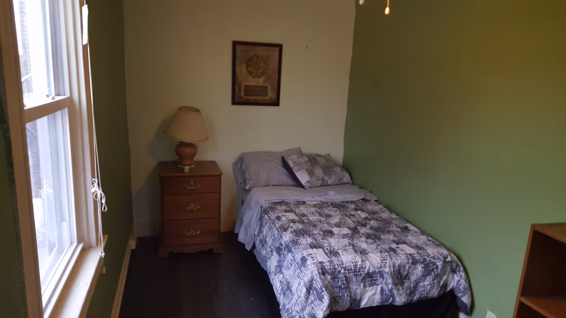 A small bedroom with a single bed covered in a gray and white patterned bedspread. Next to the bed is a wooden nightstand with three drawers and a lamp on top. A framed picture hangs on the wall above the nightstand. The walls are painted green and light beige, and there is a window on the left side letting in natural light.