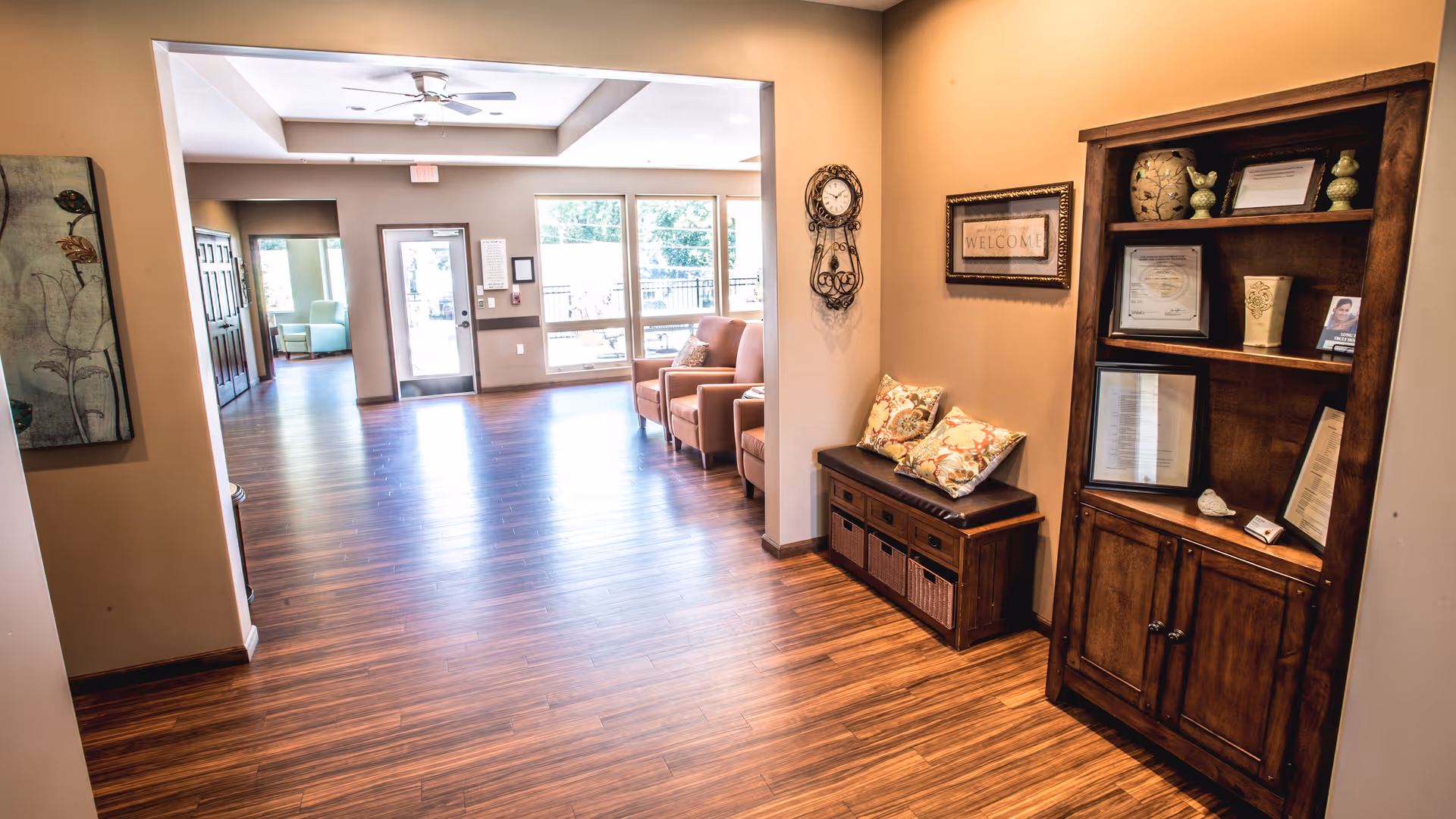 Interior view of a senior living facility lobby area with wooden flooring, a wooden bench with cushions, a wooden bookshelf with decorative items and certificates, a wall clock, and a framed welcome sign. There are large windows and a glass door letting in natural light, and a seating area with chairs is visible in the background.