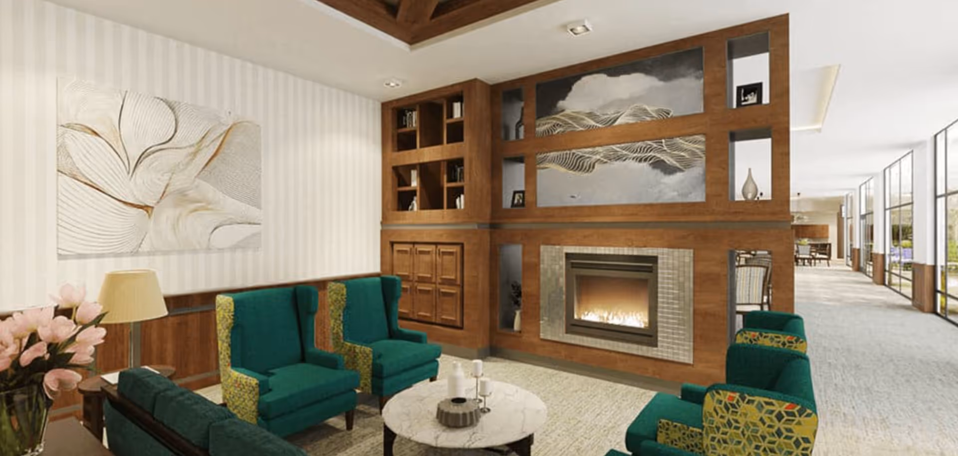 A cozy living room area in a senior living facility with green upholstered armchairs and a sofa arranged around a round marble coffee table. The room features a modern fireplace set in a wooden wall unit with built-in shelves and decorative art above it. There is a table lamp and a vase with pink flowers on a side table, and large windows along the hallway let in natural light.