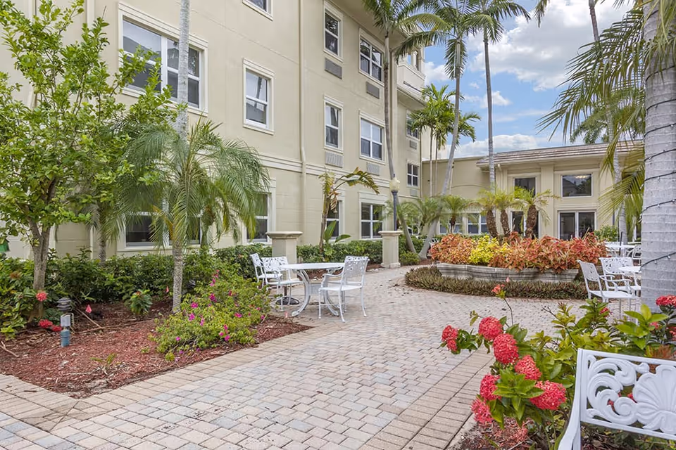 Outdoor courtyard with white patio tables, palm trees, flowering shrubs, and the beige exterior of a multi-story building.