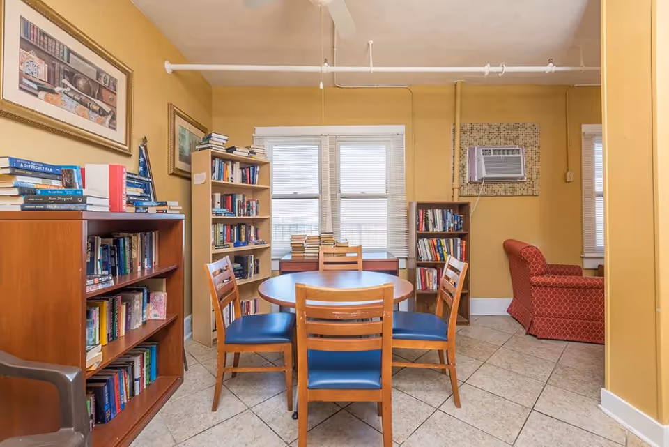 A cozy reading and sitting area in a senior living facility with yellow walls, tiled floor, and a ceiling fan. The room features multiple wooden bookshelves filled with books, a round wooden table with four wooden chairs that have blue cushions, and a red upholstered armchair. There is a window air conditioning unit installed on the wall and two windows with blinds letting in natural light.