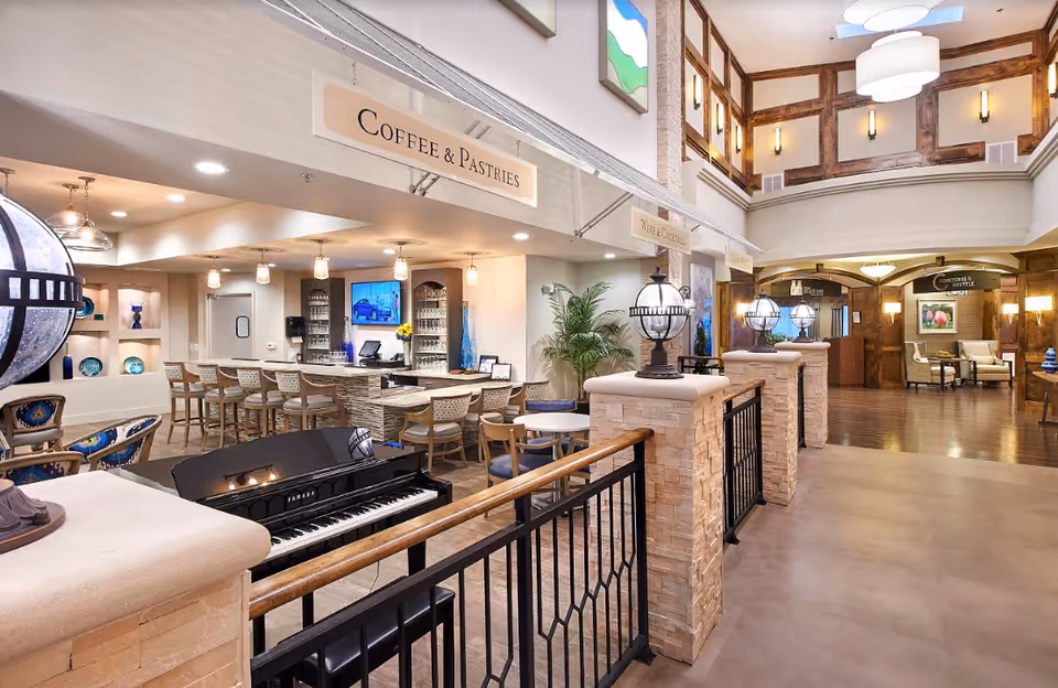 Interior view of a senior living facility featuring a coffee and pastries bar with high chairs, a black Yamaha piano in the foreground, decorative lighting fixtures, and a seating area in the background. The space has a warm, inviting atmosphere with wood accents and modern decor.