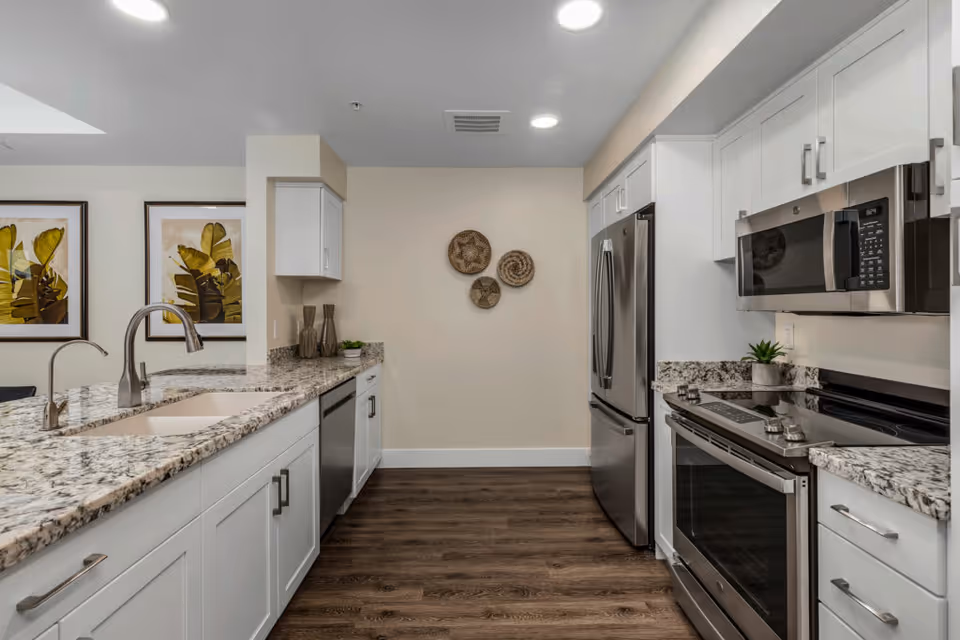 Modern kitchen with white cabinets, granite countertops, stainless steel appliances including a refrigerator, microwave, and stove. There is a double sink with a curved faucet on the left countertop. The floor is dark wood, and the wall at the end has three decorative woven baskets. Two framed botanical prints are visible on the left wall.
