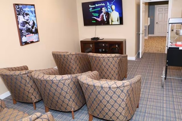 A small common area with six patterned armchairs arranged in two rows facing a wall-mounted TV. Below the TV is a wooden cabinet. On the left wall, there is a framed movie poster. The floor is carpeted with a plaid pattern, and a hallway is visible in the background.