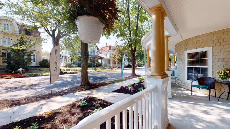 View from a porch with white railing and golden columns overlooking a street lined with trees and houses. There are flower beds along the sidewalk and a hanging white planter with green and red foliage. Two flags on the opposite side of the street read 'Assisted Living'. A black chair and a small table with a flower arrangement are on the porch.