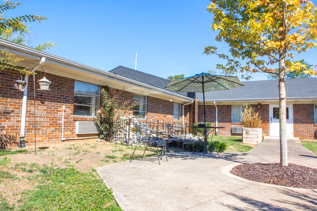 Courtyard outside a single-story brick rehab center with patio tables, an umbrella, a walkway and a tree.