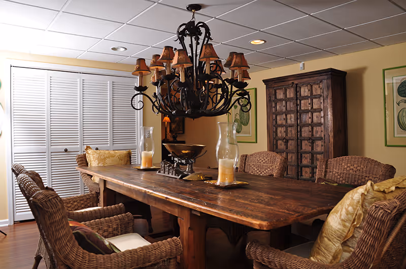 A cozy dining room with a large rustic wooden table surrounded by wicker chairs with cushions. The table is decorated with two large glass candle holders and a decorative bowl. A large ornate chandelier with multiple small lampshades hangs above the table. The room has light yellow walls, framed botanical prints, a wooden cabinet, and white louvered closet doors.