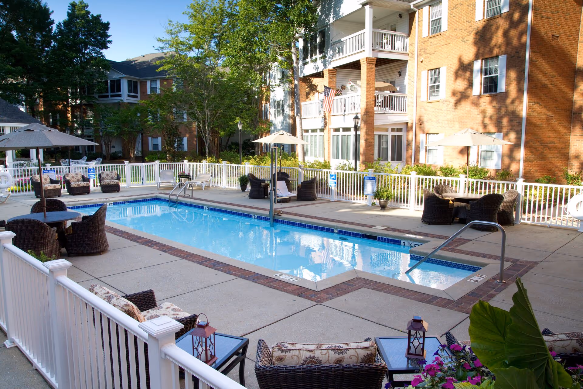 Outdoor swimming pool and patio area with wicker lounge chairs, tables, umbrellas, and a brick apartment building in the background.