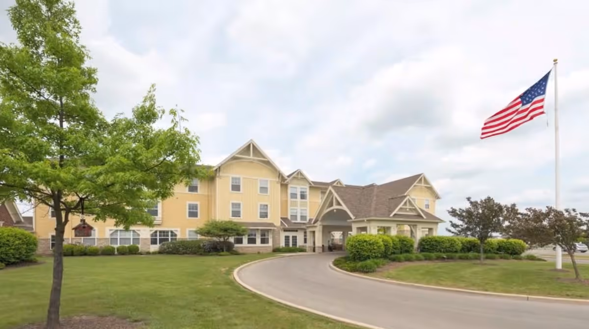 Front exterior of a yellow senior living building with a circular driveway, landscaped lawns, and an American flag flying on a flagpole.