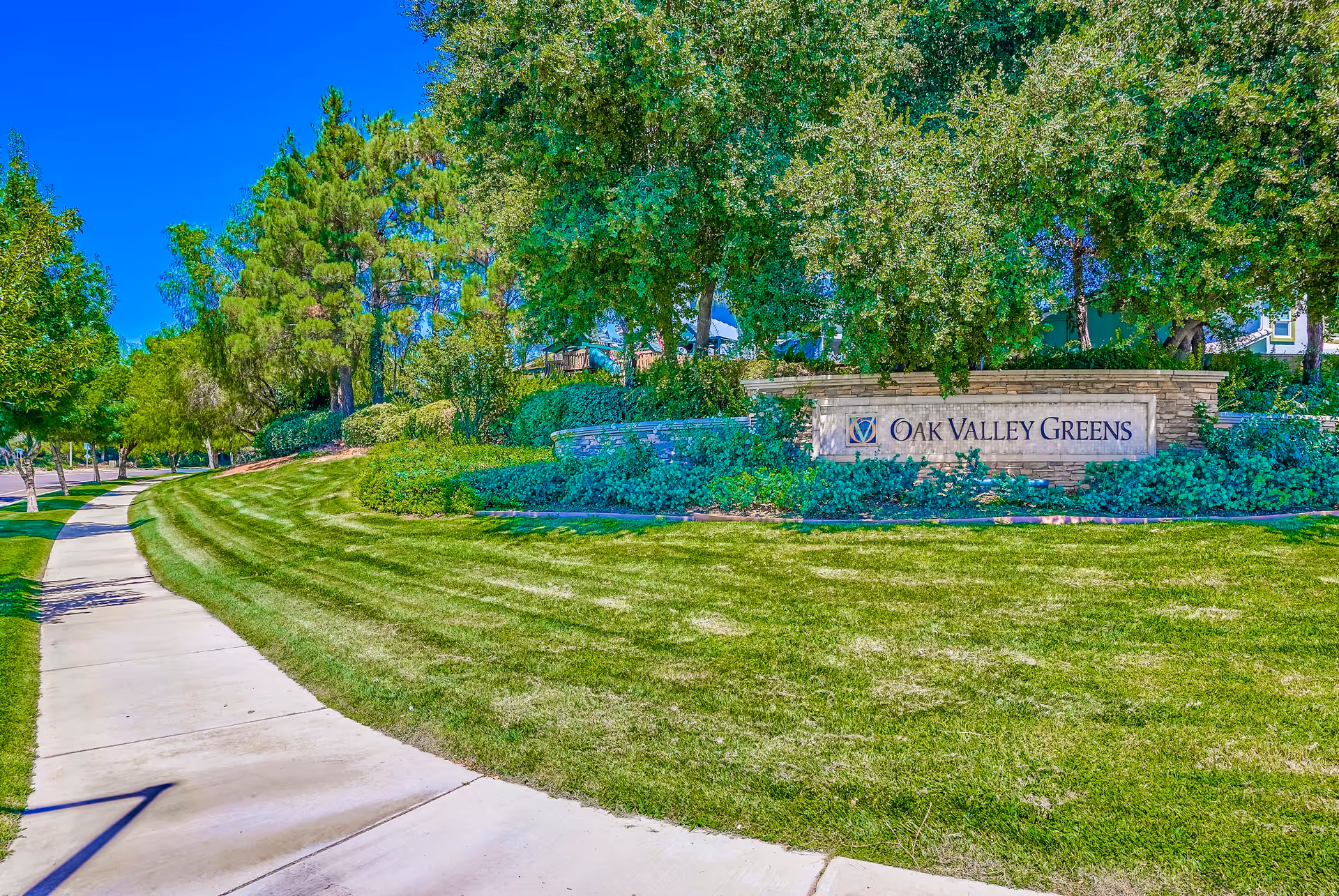 Sidewalk and manicured lawn with a stone entrance sign reading 'Oak Valley Greens' surrounded by trees and shrubs.