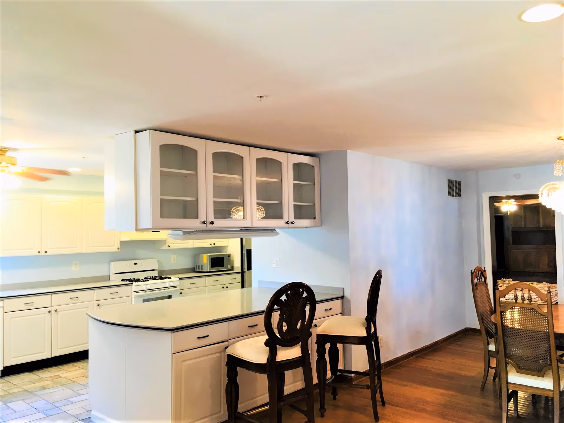 Interior view of a senior living facility kitchen and dining area. The kitchen features white cabinets, a stove, microwave, and a countertop with two wooden bar stools. Adjacent to the kitchen is a dining area with a wooden dining table and chairs, and a chandelier hanging above.