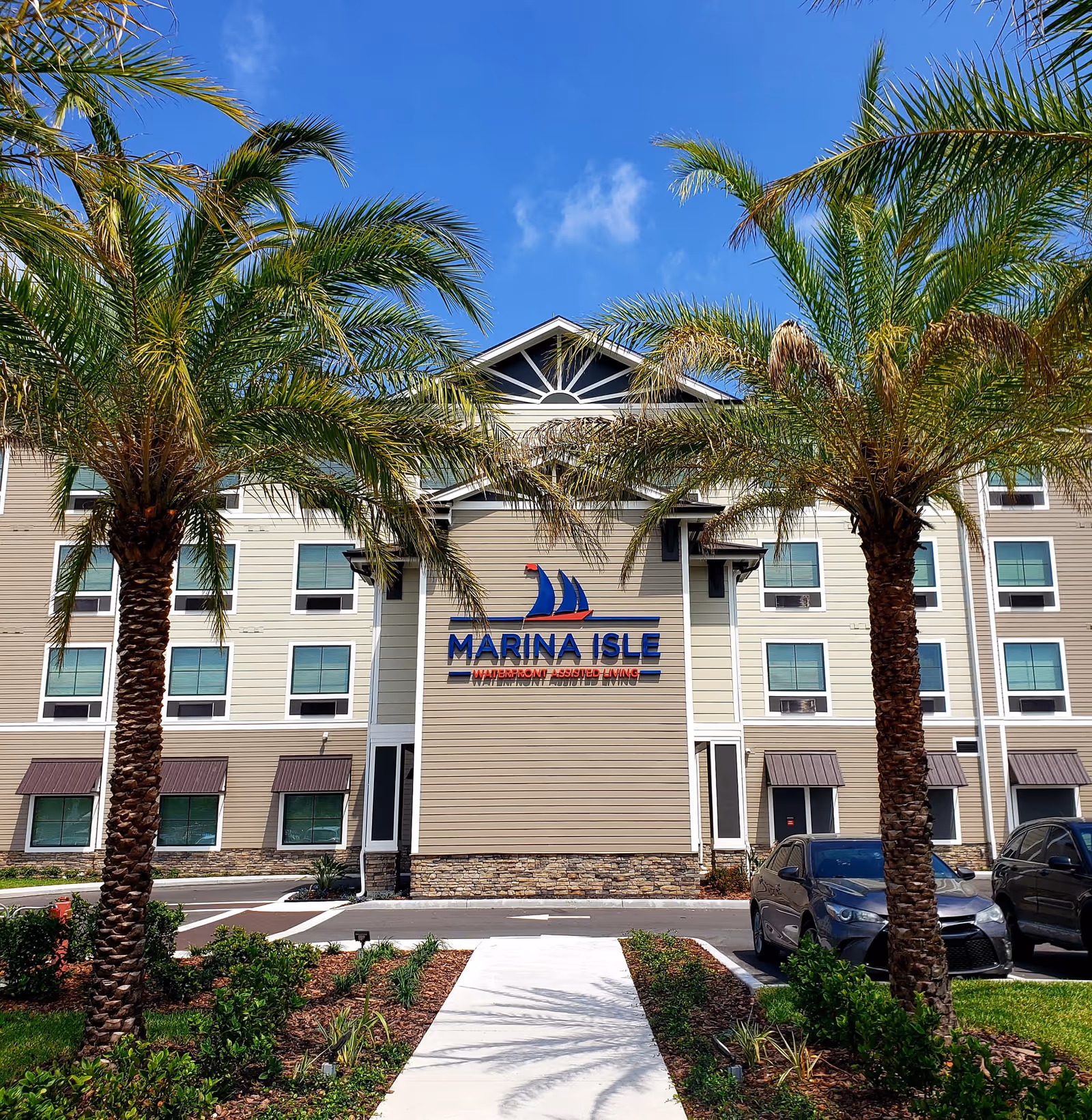 Exterior front view of Marina Isle Waterfront Assisted Living facility with palm trees, a clear blue sky, and parked cars in front.