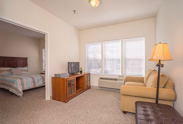 Small living room with a sofa, TV stand, floor lamp and windows, with a bedroom visible through an open doorway.