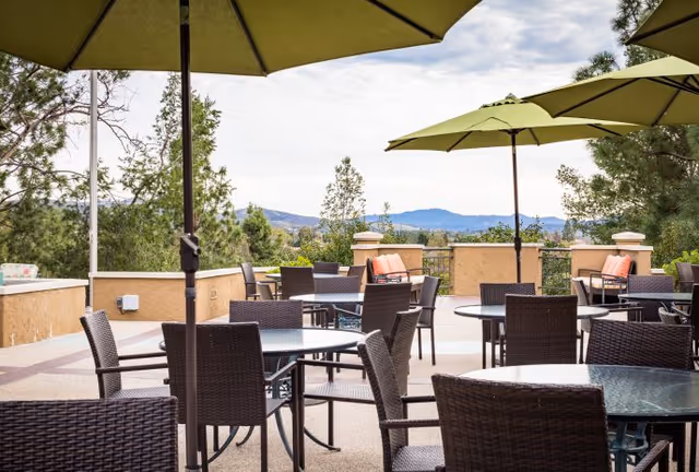Outdoor patio area with multiple round glass tables and wicker chairs under large green umbrellas, surrounded by trees and a scenic view of distant mountains under a partly cloudy sky.