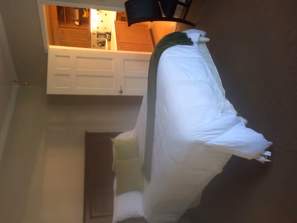 A bedroom with a neatly made bed featuring white bedding and two green pillows, a green throw blanket draped across the foot of the bed, a wooden headboard, and an open door leading to a kitchen with wooden cabinets and a countertop. A dark chair is partially visible near the door.
