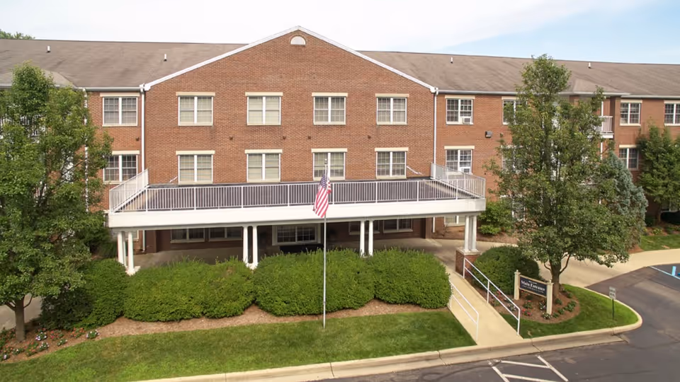 Front exterior of a three-story brick senior living building with a covered entrance, balcony, American flag, and landscaped shrubs.