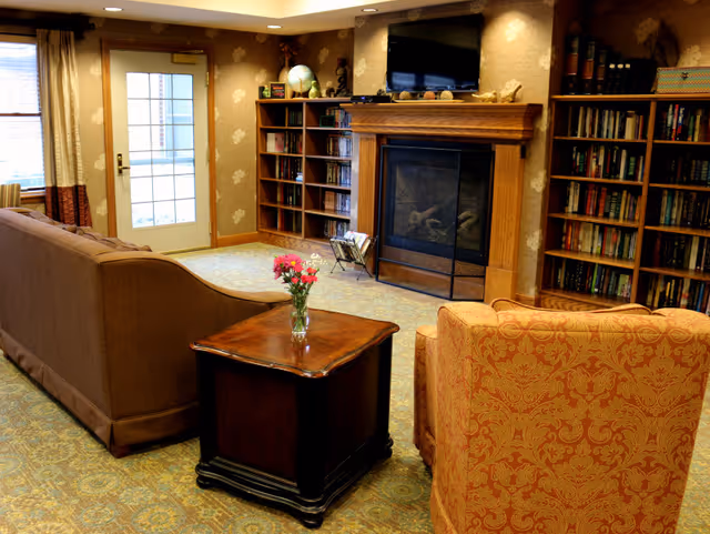 Cozy common living room with a sofa and armchair facing a fireplace and TV, with bookshelves and a small table with flowers.