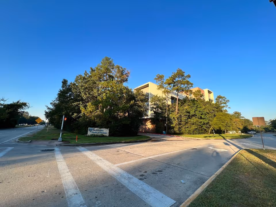 Exterior view of a multi-story building partially obscured by trees under a clear blue sky, with a street intersection and a sign for Village at Woodlands Waterway in the foreground.