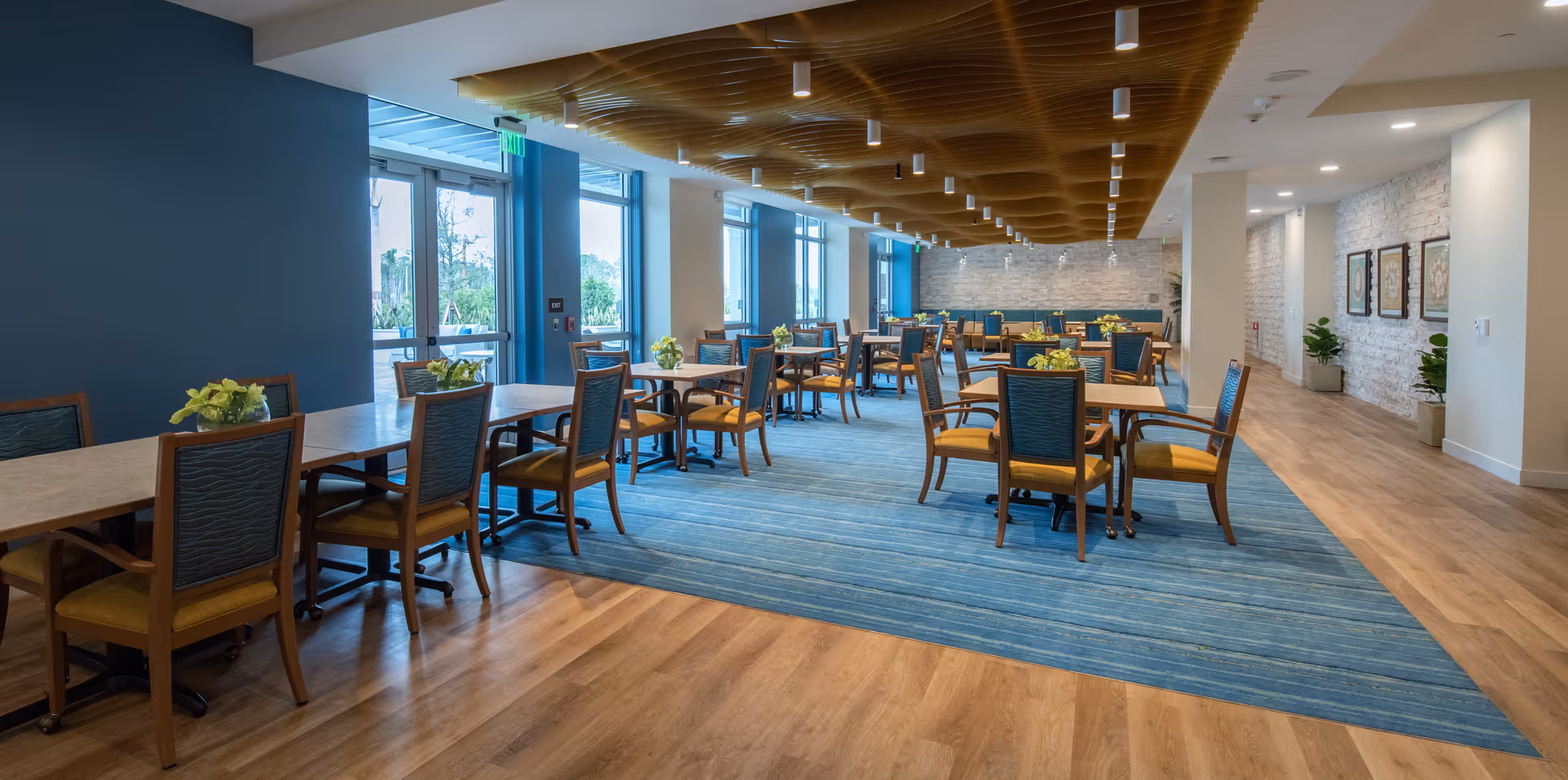 A spacious dining room with multiple wooden tables and chairs arranged neatly on a blue carpet. The room features large windows allowing natural light to fill the space, a wooden ceiling design, and decorative plants and framed artwork along the walls.