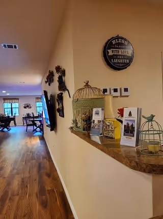 Interior hallway of a senior living facility with wooden flooring and beige walls. On the right side, a countertop displays decorative items including a birdcage, brochures, and a small bird figurine. The wall above the countertop has a round decorative sign that reads 'Bless this house with love and laughter.' In the background, there is a dining area with tables and chairs near windows.