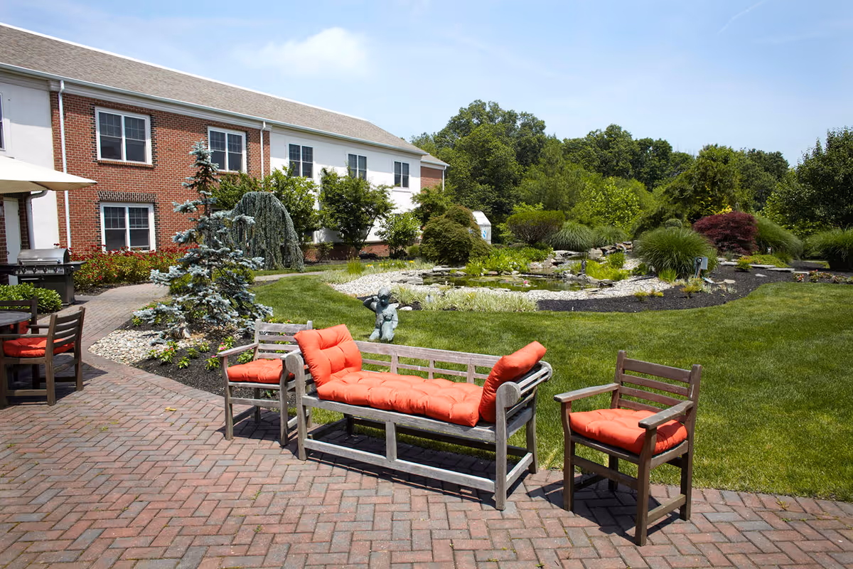 Outdoor patio area with wooden chairs and a bench featuring red cushions, set on a brick-paved surface. In the background, there is a well-maintained garden with green grass, various shrubs, a small pond, and a two-story brick and white building under a clear blue sky.