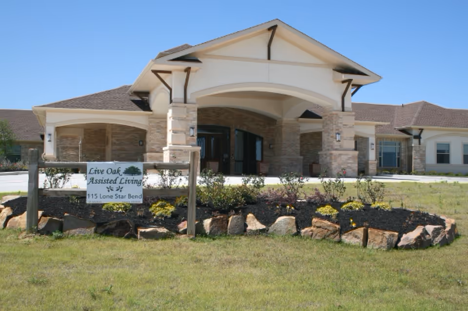 Front exterior view of a single-story assisted living facility with a covered entrance, stone pillars, and a landscaped garden with rocks and flowers in front. A sign in the garden reads 'Live Oak Assisted Living 115 Lone Star Bend'.