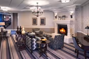 A cozy and elegantly decorated living room area with patterned armchairs, a sofa, a coffee table, and a lit fireplace. The room features striped carpeting, framed artwork on the walls, a chandelier, and decorative items on tables and the mantelpiece.