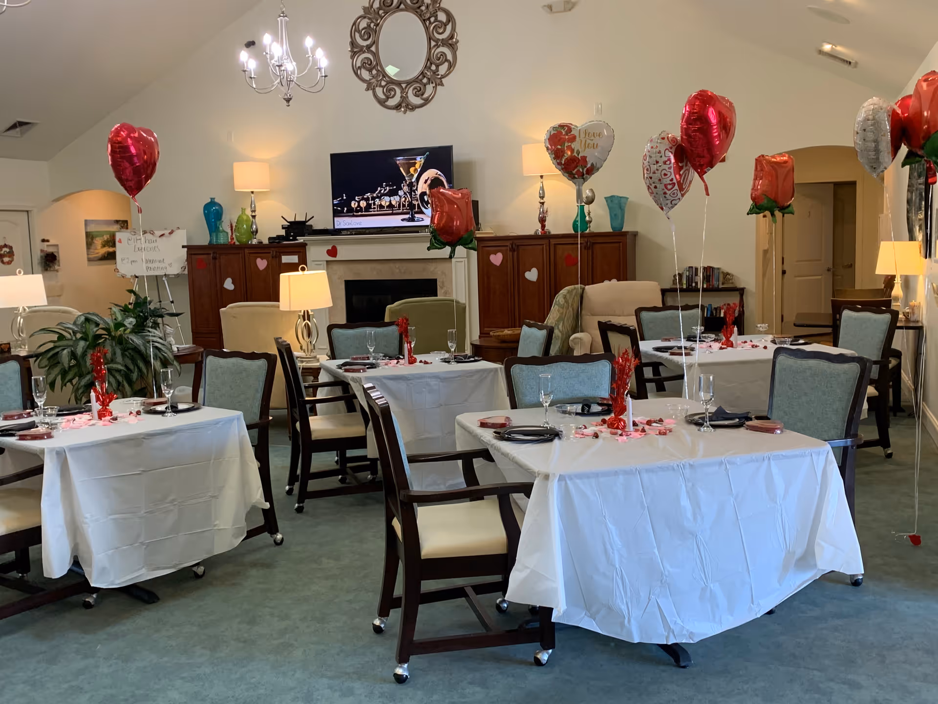 A decorated dining room in an assisted living facility with tables covered in white tablecloths, set with plates, glasses, and napkins. Red and heart-shaped balloons are tied to the chairs, and red decorations are scattered on the tables. The room has a fireplace with a TV above it, lamps, and a mirror on the wall. Comfortable chairs and cabinets are visible in the background.