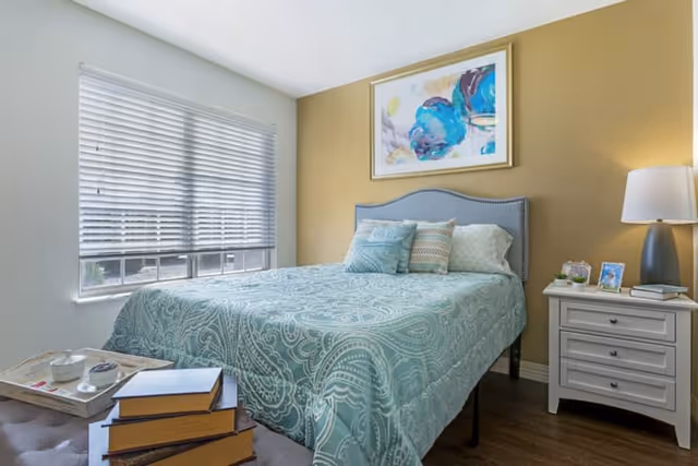 A bedroom with a bed covered in a teal patterned bedspread and several pillows. There is a nightstand with a lamp, a small plant, and framed photos. A window with white blinds lets in natural light. A colorful abstract painting hangs on the wall above the bed.