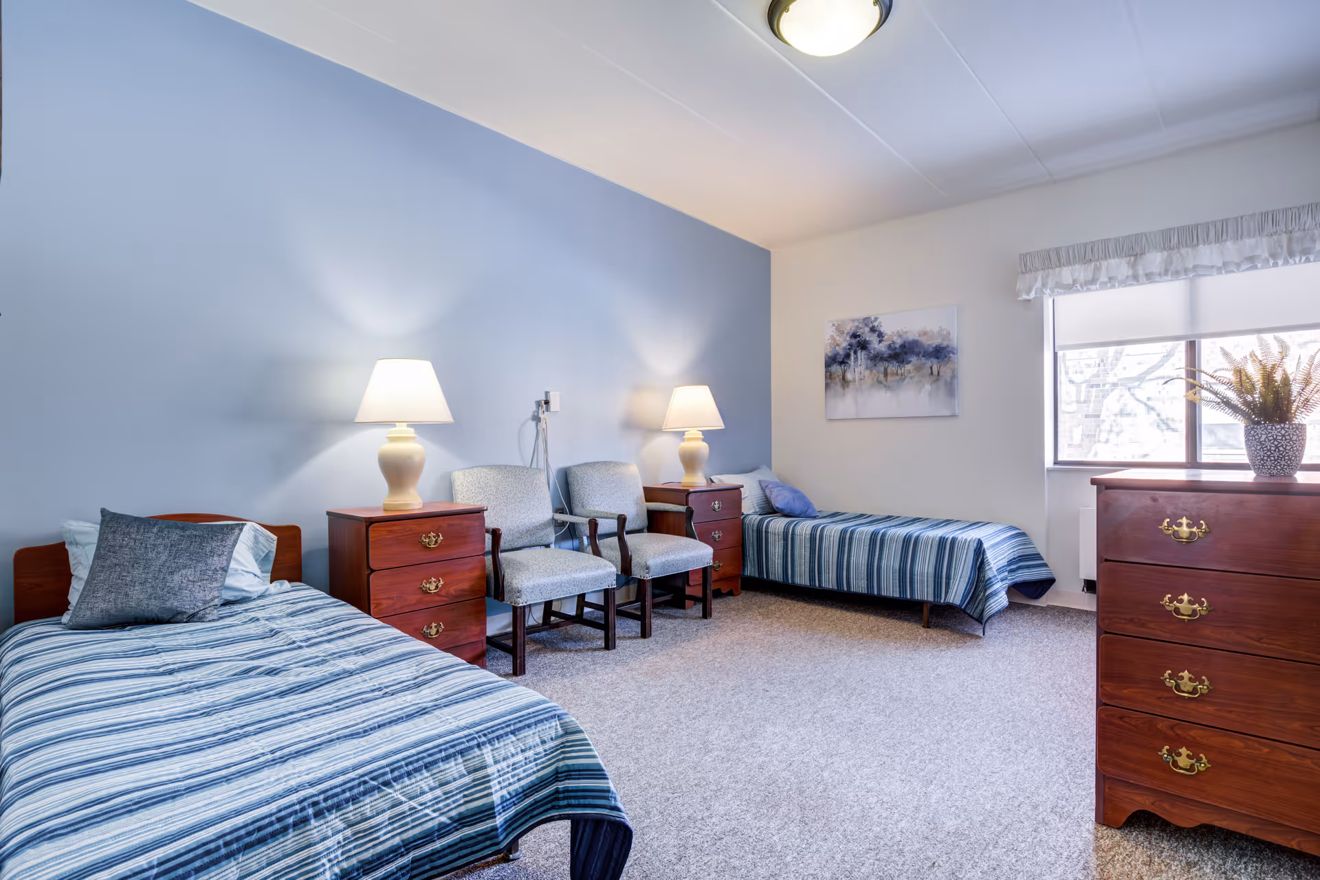 A bright and tidy bedroom with two single beds covered in blue striped bedding. Between the beds are two wooden nightstands with matching lamps. Two upholstered chairs are placed side by side against a light blue accent wall. A window with a white valance lets in natural light, and a wooden dresser with a potted plant sits nearby. A framed artwork hangs on the wall above one of the beds.