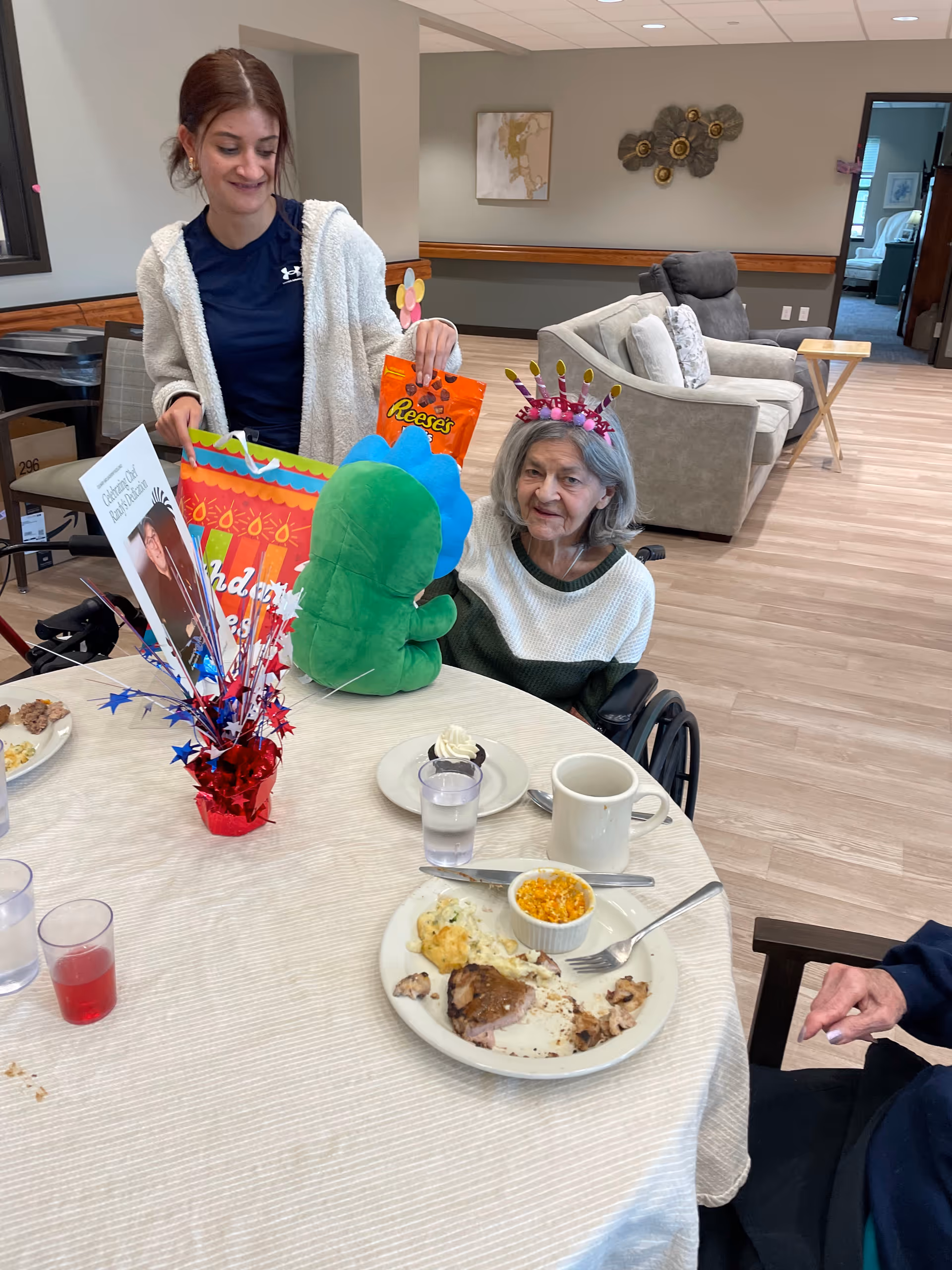An elderly woman wearing a birthday crown sits in a wheelchair at a dining table with a partially eaten meal, a dessert, and drinks. A younger woman stands beside her holding a gift bag and a Reese's candy package. The room has comfortable seating and light wood flooring.