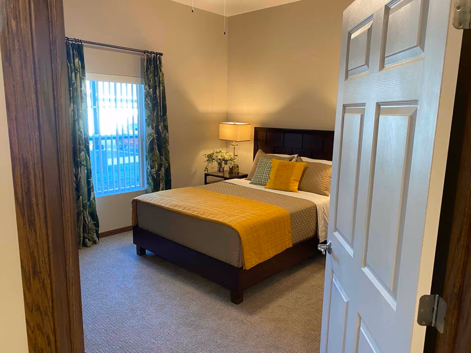 View through an open door into a bedroom with a neatly made bed featuring a yellow and gray bedspread and pillows. A bedside table with a lamp and a vase of flowers is next to the bed. A window with vertical blinds and green patterned curtains is on the left wall.