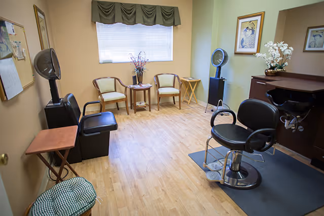 Interior salon room with a barber chair, hooded hair dryers, waiting chairs, and a window.