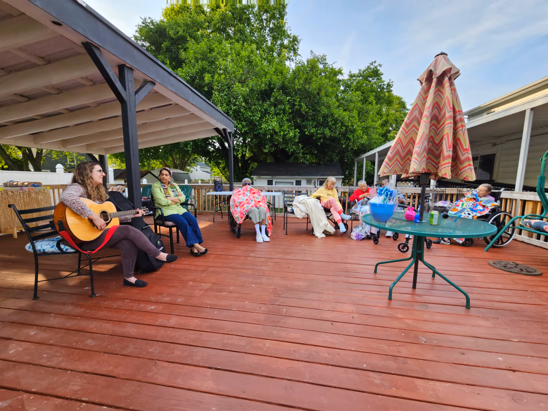 A group of elderly people sitting on a wooden deck outdoors, some in wheelchairs and others on chairs, with a woman playing guitar. The deck has a covered area on the left and a round glass table with a closed umbrella on the right. Trees and a small building are visible in the background.