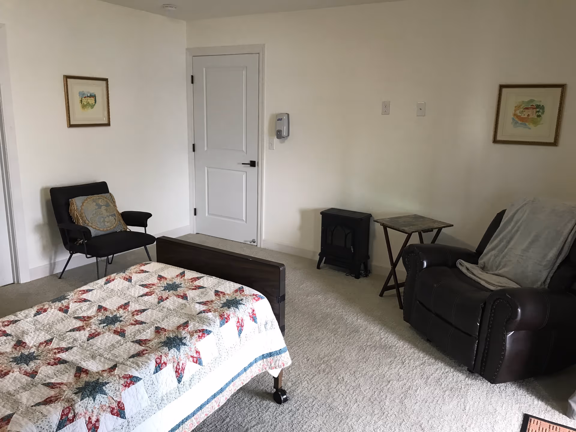 A bedroom with a bed covered in a patterned quilt, a black armchair with a decorative pillow, a brown recliner with a gray blanket draped over it, a small side table, a small black heater, and two framed pictures hanging on the walls.