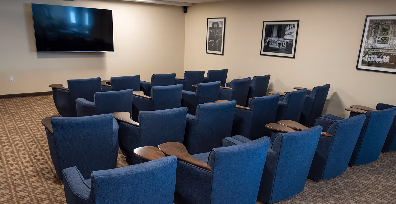 A small theater-style room with multiple rows of blue upholstered chairs, each equipped with wooden armrest trays, facing a large flat-screen TV mounted on a beige wall. The room has patterned carpet flooring and three black-and-white framed photographs hanging on the walls.