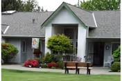 Exterior view of a single-story building with a gray roof and white siding, featuring a small covered entrance with a bench and landscaped bushes and flowers in front.