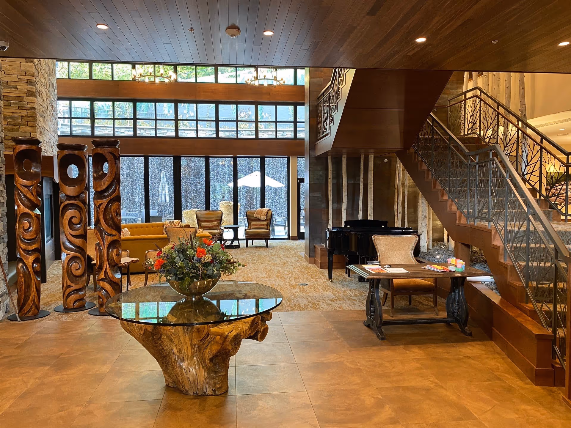 Spacious and warmly lit interior of a senior living facility featuring a wooden table with a glass top and a floral arrangement in the center. To the left, there are three tall carved wooden sculptures. In the background, there are comfortable chairs and sofas arranged near large windows with a view of an outdoor patio. A grand piano is positioned near a staircase with decorative railings on the right side.
