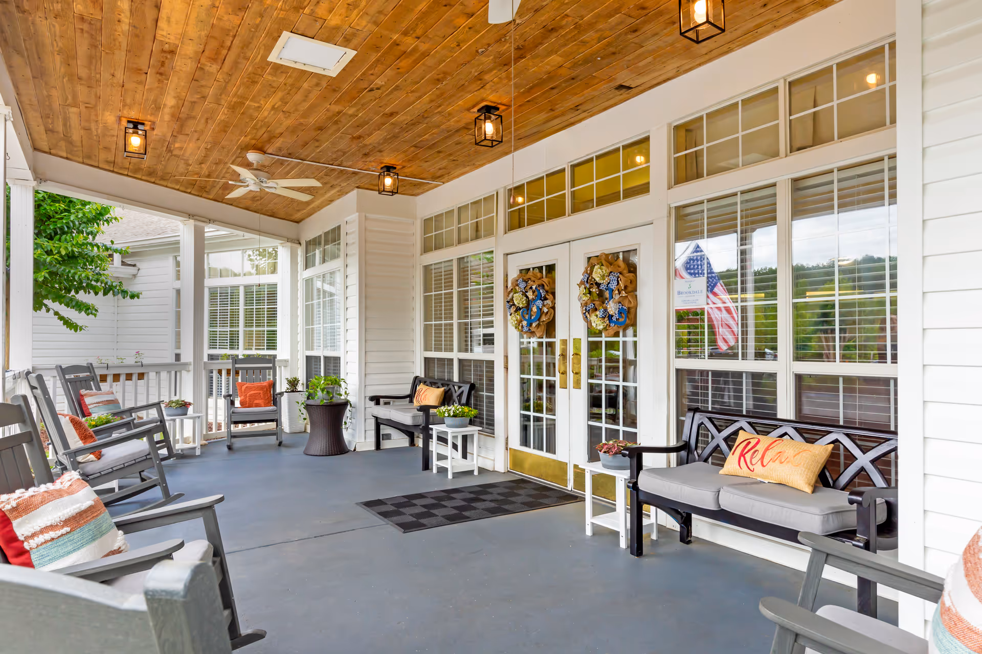 Covered porch area at Brookdale Lexington with wooden ceiling, ceiling fan, and hanging lights. The porch has multiple rocking chairs and benches with cushions and pillows, some with the word 'Relax'. There are decorative wreaths on the glass double doors and potted plants on small tables. An American flag is visible through the window.
