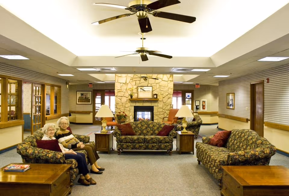 A cozy assisted living facility common area with floral patterned sofas and armchairs arranged around a stone fireplace. Two elderly women are seated on one of the sofas. The room has ceiling fans, table lamps, framed pictures on the walls, and a carpeted floor.