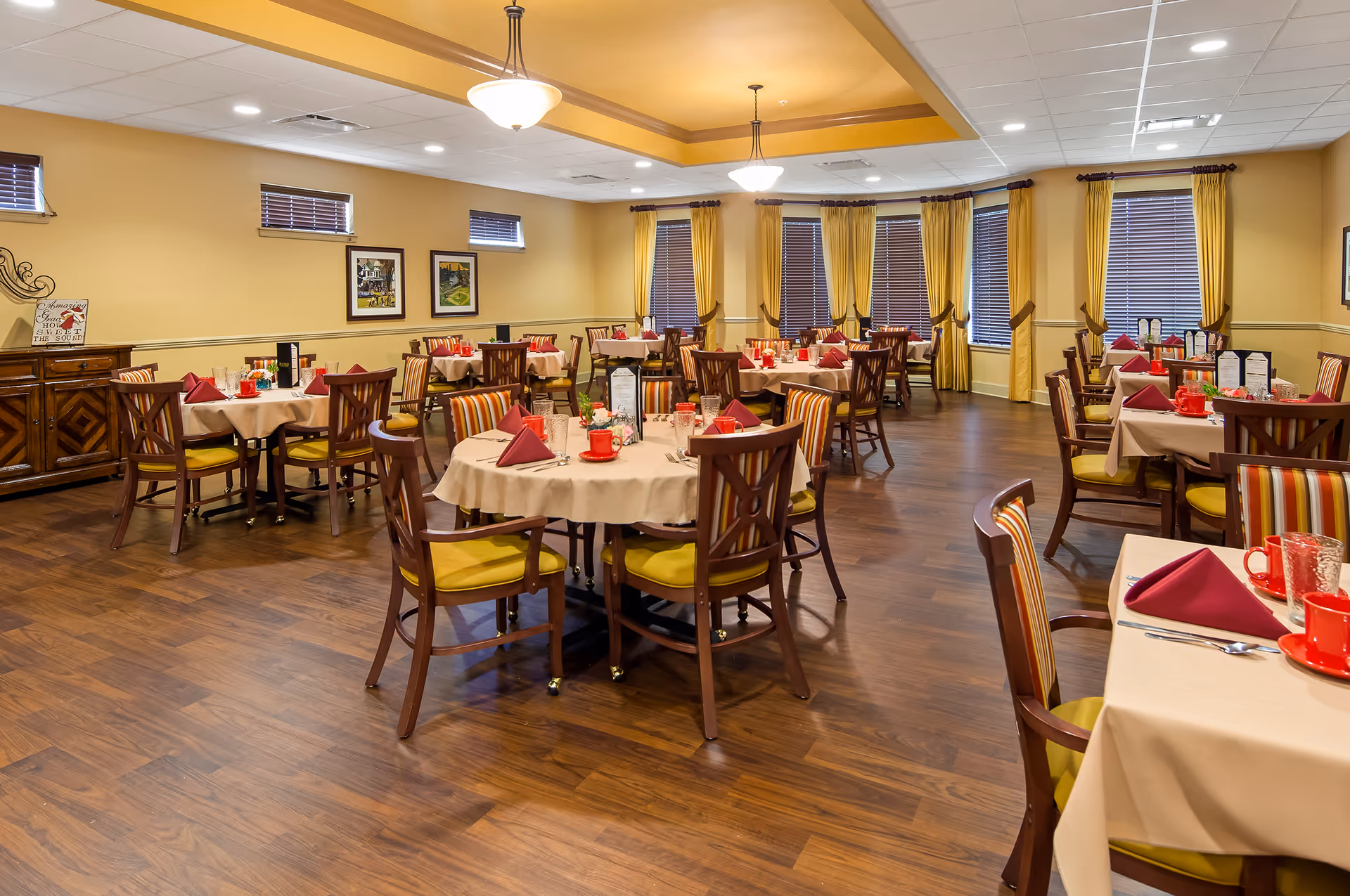A spacious dining room with multiple round tables covered with beige tablecloths, each set with red napkins, red cups, glasses, and silverware. The chairs have wooden frames with yellow and striped upholstery. The room has wooden flooring, yellow walls, several windows with brown blinds and yellow curtains, and ceiling lights providing warm illumination.