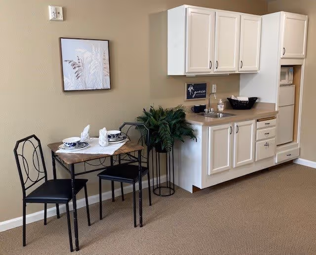 Small dining area with a wooden table set for two with plates, bowls, and napkins, next to a kitchenette with white cabinets, a countertop with a basket and cups, and a potted plant in the corner.