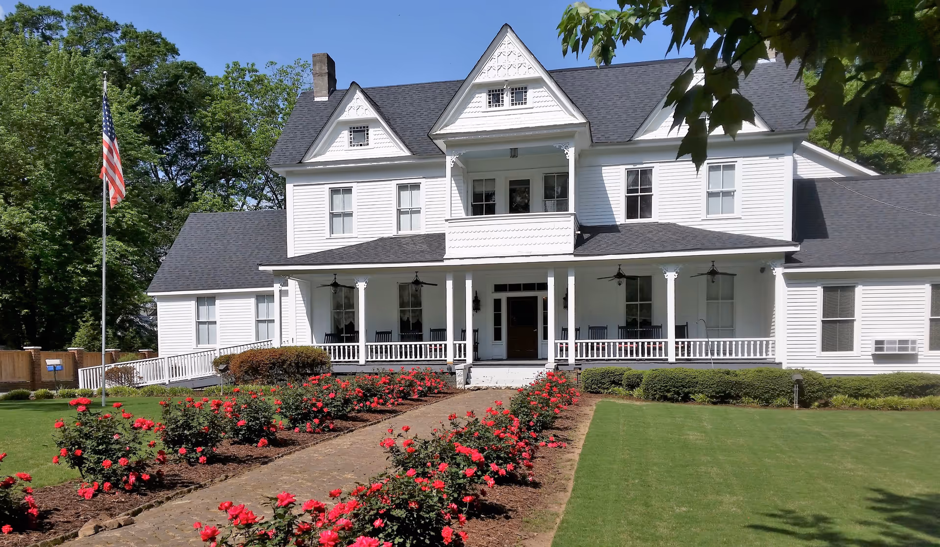 Front exterior view of a large white two-story house with a dark shingled roof, a porch with rocking chairs, a balcony, and a garden with red flowers lining a stone pathway leading to the entrance. An American flag is on a flagpole to the left, and trees surround the property.