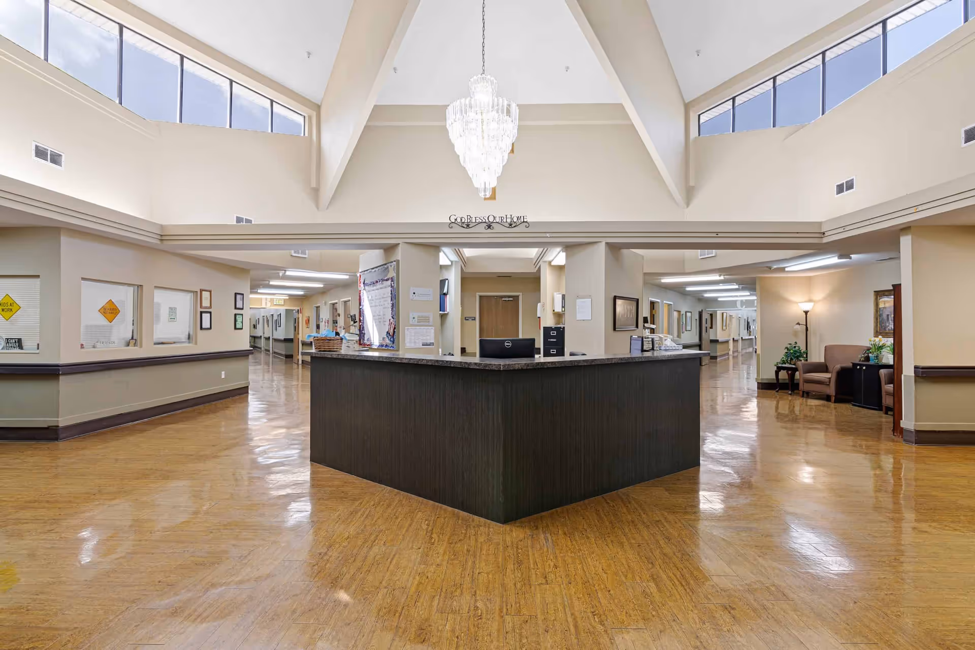 Interior view of a nursing and rehab facility reception area with a large dark wood reception desk in the center, polished wooden floors, beige walls, high vaulted ceilings with triangular windows, a chandelier hanging from the ceiling, and hallways extending on both sides. There are some chairs and plants in a seating area to the right.