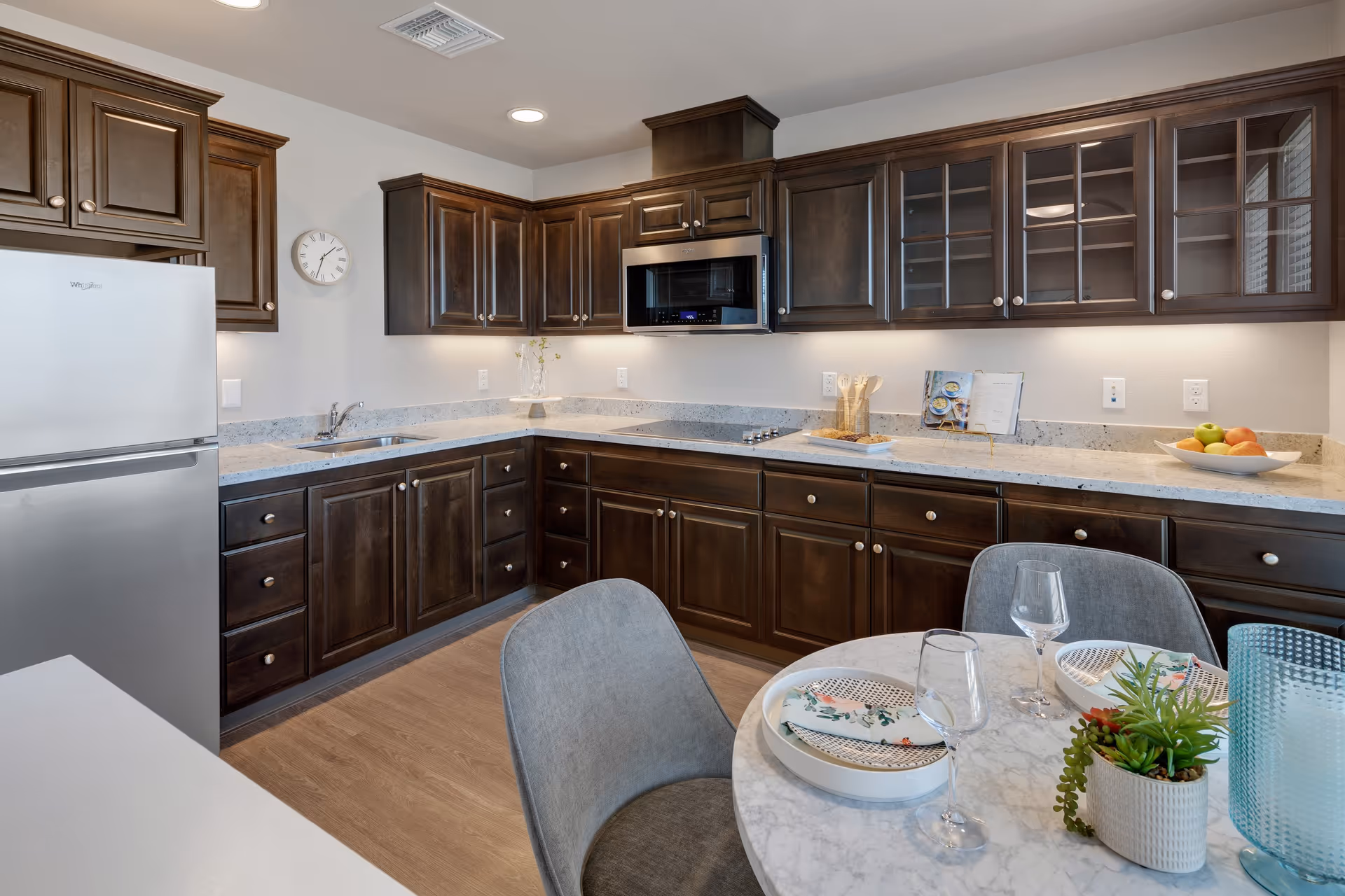 Modern kitchen with dark wooden cabinets, a white refrigerator, a built-in microwave above the stove, and a marble countertop. A round dining table with two gray chairs is set with plates, napkins, wine glasses, and a small plant. The kitchen has under-cabinet lighting and a wall clock.