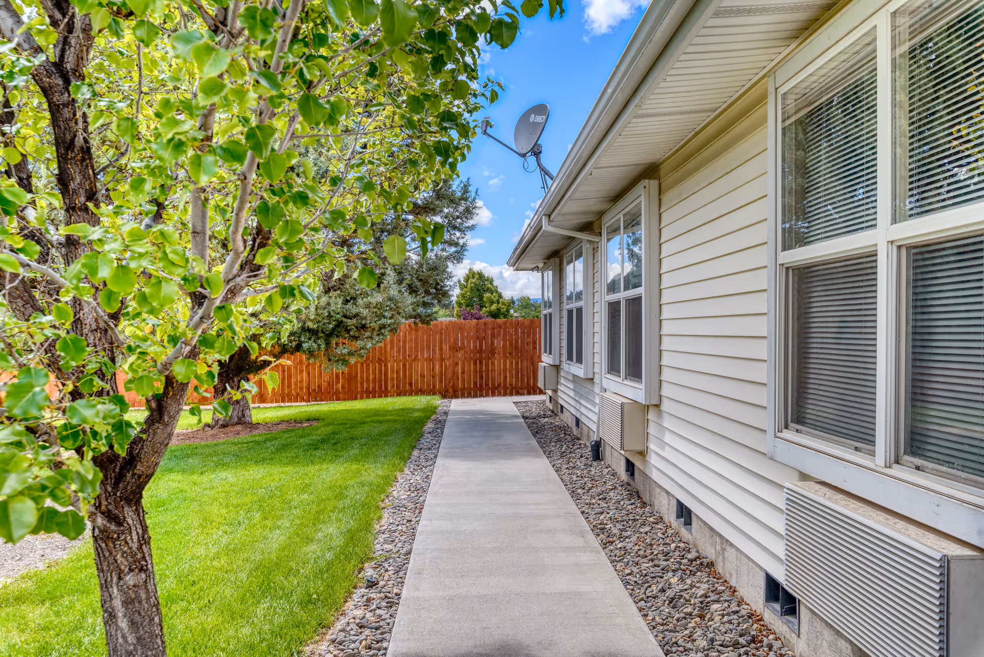 A concrete walkway runs alongside the exterior of a beige building with several windows. The building is bordered by a strip of rocks, and a wooden fence is visible at the end of the walkway. On the left side, there is a green lawn with trees and shrubs under a partly cloudy blue sky.