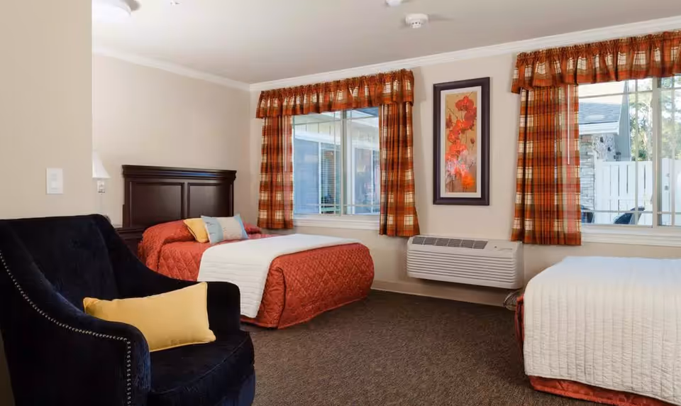 A cozy bedroom with two beds, one with a red quilt and the other with a white quilt. Both beds have pillows and are positioned near windows with orange plaid curtains. A dark blue armchair with a yellow pillow is placed in the corner. A framed floral artwork hangs on the wall above a wall-mounted air conditioning unit.