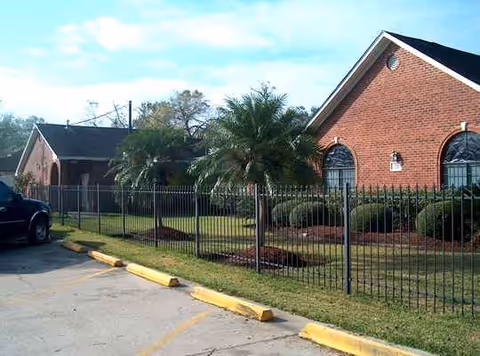 Brick assisted living facility exterior with a metal fence, palm trees, and a parking lot.