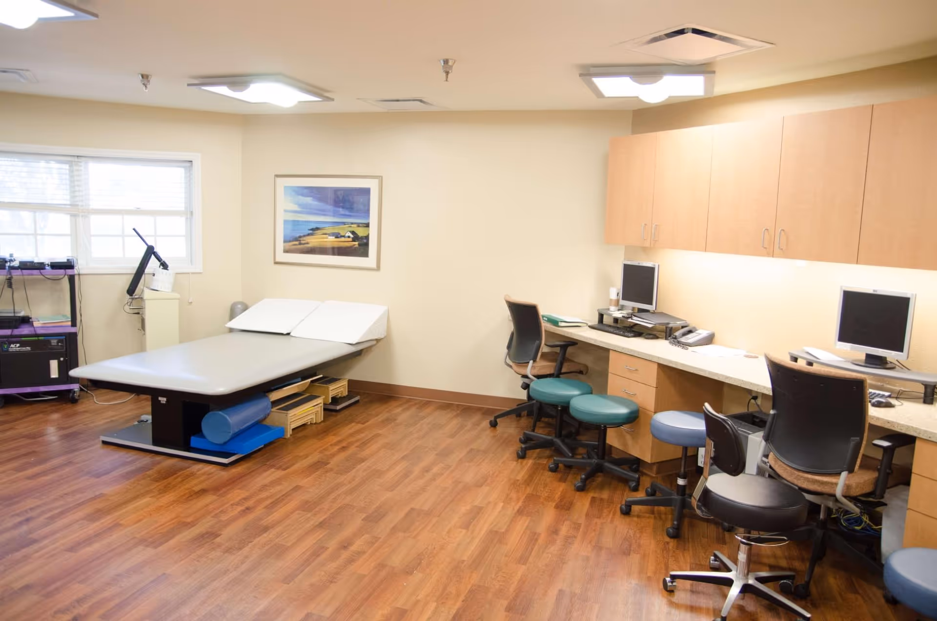 A bright therapy/exam room with an adjustable treatment table, rolling stools and desk workstations with computers.